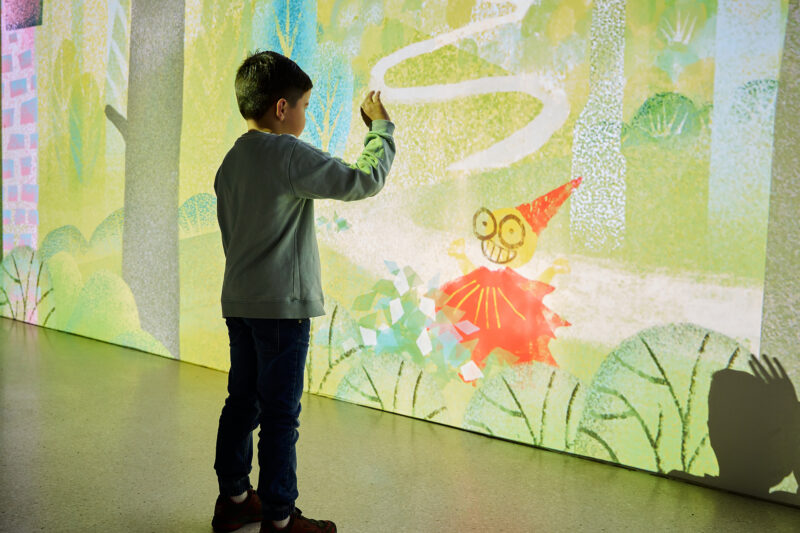 05 | IMMERGRUEN | GRIMMWELT Kassel | Foto: Nicolas Wefers Ein Junge spielt mit einer gestempelten Firgur im virtuellen Märchenwald an der Wand.