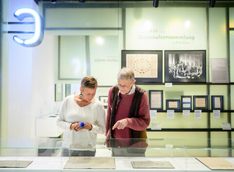 C | CASSEL | Foto: Sascha Mannel Ein älterer Herr zeigt seiner Begleitung Ausstellungstücke in einer Vitrine im Bereich CASSEL.