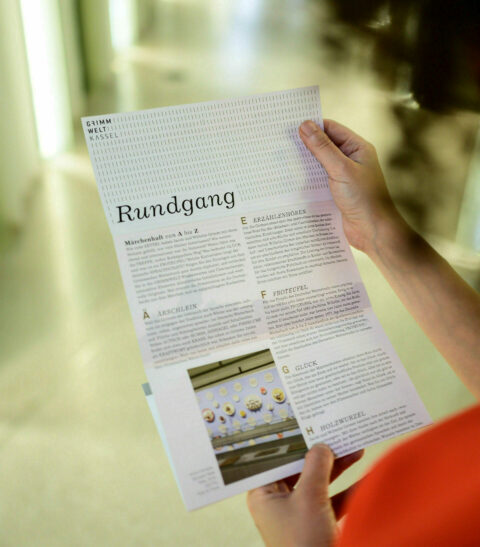 GRIMMWELT Kassel | Permanent Exhibition | Flyer "Tour guide" | Photo: Sascha Mannel A person holds the flyer "Tour guide" in hands.