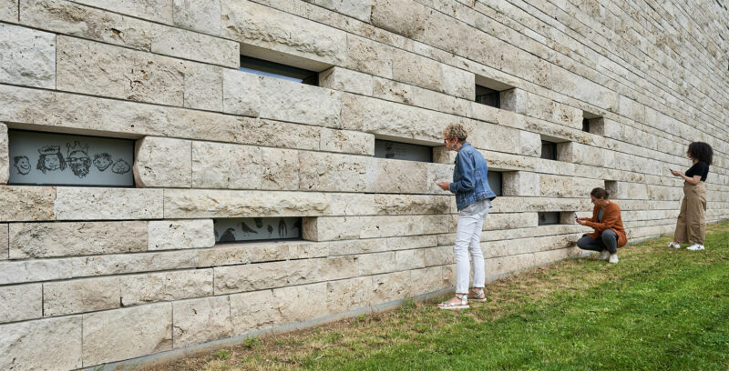 DrachenFels | FindeLohn | Foto: Nikolaus Frank Drei Frauen betrachten Zeichnungen in kleinen Nischenfenstern auf der Rückseite der GRIMMWELT.