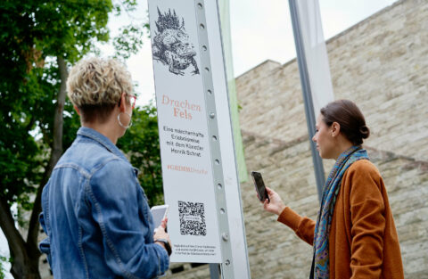 DrachenFels | Start der Reise | Foto: Nikolaus Frank 2 Frauen scannen den QR-Code, um der DrachenFels-Reise um die Grimmwelt herum zu folgen.
