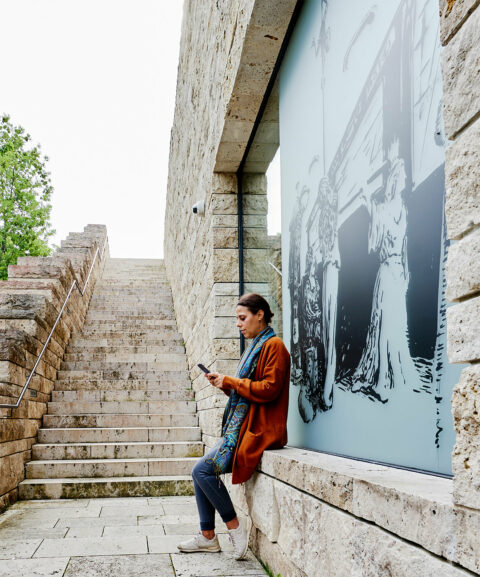 DrachenFels | ReisePlan | Foto: Nikolaus Frank Frau sitzt vor der Scheibe der Station ReisePlan der Erlebnisreise DrachenFels und betrachtet einen digitalen Inhalt auf ihrem Smartphone.