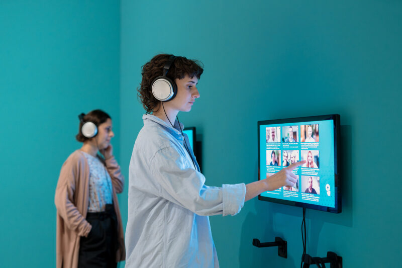 Ausstellung ICH, DAS TIER | Interviewstation | © GRIMMWELT Kassel, Foto Nicolas Wefers 2 Frauen stehen mit Kopfhöhrern an der Interviewstation