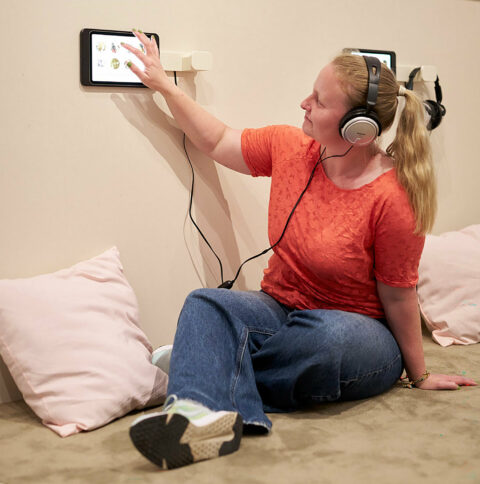 Sonderausstellung ZWISCHEN GESTALTEN. HELGA GEBERT | »Leseecke« | Foto: Nicolas Wefers Ausstellungsansicht: Frau sitzt in der Leseecke auf dem Boden und hört Märchen