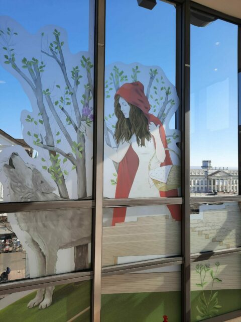 GRIMMWELT zu Gast in der GALERIA | Rotkäppchen-Illustration am Fenster Blick auf eine Installation von Rotkäppchen, die an den Fensterscheiben von GALERIA befestigt wurde.