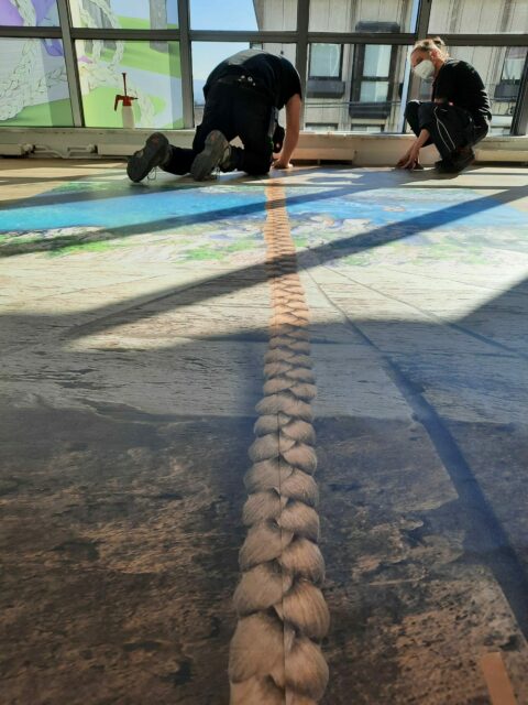 GRIMMWELT zu Gast in der GALERIA | Rapunzelzopf bei GALERIA Die Illustration eines blonden, langen Zopfes für Rapunzel wird auf dem Boden angebracht.